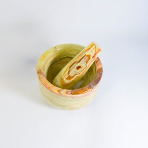 May include: A green, yellow, and brown onyx mortar and pestle set. The mortar is a round bowl with a matching pestle resting inside. The natural stone features a striped pattern with variations of color.