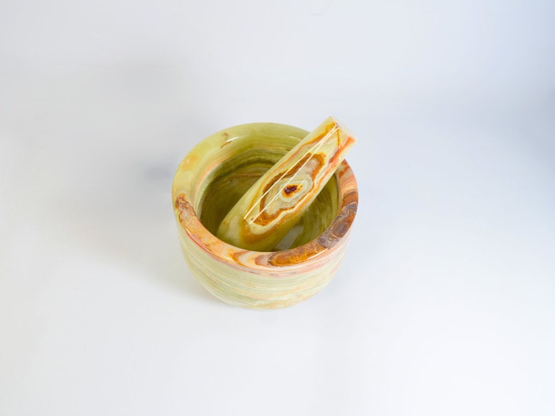 May include: A green, yellow, and brown onyx mortar and pestle set. The mortar is a round bowl with a matching pestle resting inside. The natural stone features a striped pattern with variations of color.