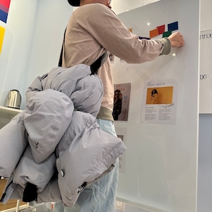 May include: A person wearing a light pink long-sleeved shirt, a black baseball cap, and light blue jeans is standing in front of a white board. The person is holding a light blue puffer jacket with a black strap. The white board has a list of local events written on it, including "June 27 - Pride Toronto!" and "Reese Miller".