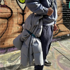 May include: A grey puffer jacket with a black zip and a teal strap. The jacket is folded and being carried over the shoulder of a person.