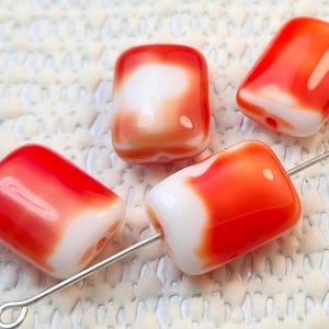 May include: Four rectangular beads with rounded edges, featuring a vibrant red and white color scheme. The beads are strung on a silver-colored wire, suggesting their use in jewelry making or crafting. The beads have a glossy finish.