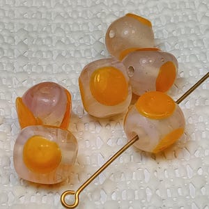 May include: Five glass beads with a translucent white base and opaque yellow circles. The beads are round and square shaped.