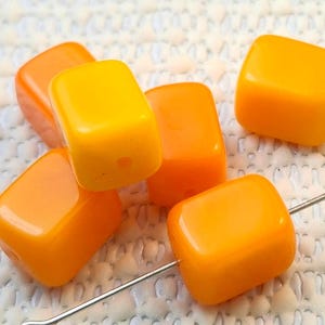 May include: Six glossy, orange and yellow cube-shaped beads are displayed. The beads are strung on a silver-colored wire, and have a hole drilled through the center. The beads are arranged on a white, textured surface.