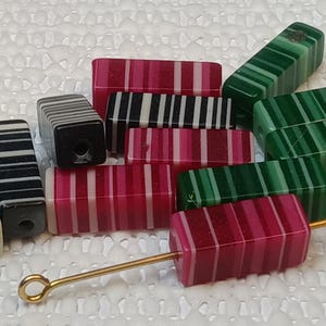 May include: Assortment of colorful striped polymer clay beads in black and white, pink and white, and green and white. The beads are rectangular and have a hole through the center. A gold metal beading needle is also included.