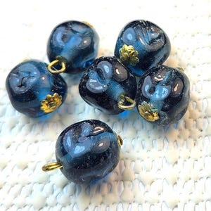 May include: Six dark blue glass beads with gold-colored accents. Each bead has a unique shape and a loop for stringing. The beads have a marbled appearance with swirls of lighter blue.