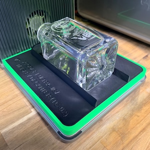 May include: A clear, rectangular glass bottle with a decorative pattern and etched design. The bottle sits on a black platform with green trim, inside a larger device. The text on the bottle is partially visible.