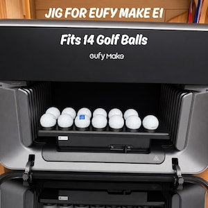 May include: A black and gray device labeled "JIG FOR EUFY MAKE E1" with the text "Fits 14 Golf Balls" and "eufy Make." The device holds 10 white golf balls on a shelf. The device has a hinged door.