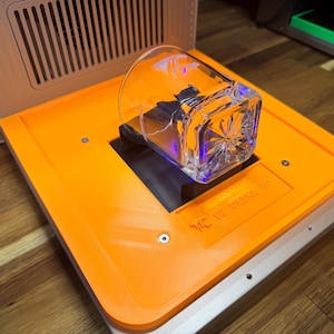 Op de afbeelding: Een helder, vierkant glas met een decoratief patroon, rustend op een oranje platform. Het platform maakt deel uit van een groter apparaat met een houten en metalen frame. Het glas wordt verlicht met blauw licht.