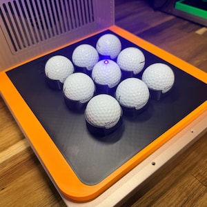 May include: A golf ball alignment device with nine white Titleist golf balls arranged on a black surface. The device has an orange border and a light source in the center. The device is on a wooden surface.