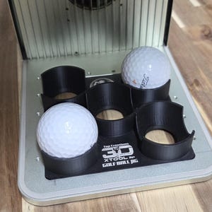 以下が含まれることがあります： 3つの白いゴルフボールが入った、シルバーと黒のゴルフボールジグ。ジグは、"3D XTOOL Golf Ball Jig"のテキストが入った黒いベースを持っています。ゴルフボールは黒いホルダーに入っています。