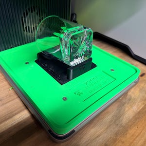 May include: A clear, square-shaped glass with a decorative pattern sits on a black stand, placed on a bright green surface. The surface has text that reads "250ml" and "MADE IN USA". The setup is inside a gray and black machine.