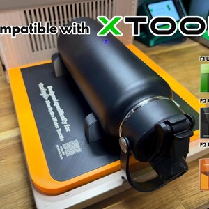 Puede incluir: Botella de agua negra sobre una base negra y naranja, con el texto "Compatible with XTOOL". La base está diseñada para una botella de agua Hydro. La imagen también muestra los modelos F1 Ultra, F2 Ultra y F2 Ultra UV.
