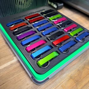 Può includere: Una vetrina piena di portachiavi colorati. I portachiavi sono in vari colori, tra cui rosso, blu, verde e nero. La vetrina ha un interno nero e un bordo verde, su una superficie di legno.