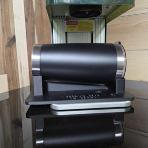 May include: A black cylindrical object with silver accents, possibly a tumbler or container, is mounted on a black base. The object is part of a larger machine with a green top. The background features wooden paneling.
