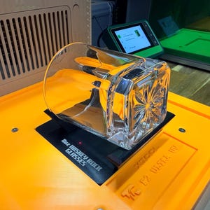 Op de afbeelding: Een helder, vierkant whiskyglas met een textuurontwerp staat op een zwarte basis. De basis staat op een feloranje oppervlak, met de tekst "8oz WHISKEY ROCK GLASSES" zichtbaar. Een groen scherm en andere apparatuur staan op de achtergrond.