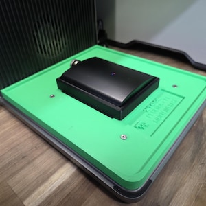 May include: A black flask with a silver cap rests on a bright green tray. The tray is set inside a larger, metallic frame. The tray has the text "PROCES... FILTER + MULTI-PASS BASE" printed on it. The background includes a dark, ribbed structure.