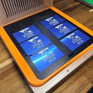 May include: A jig holder in orange and black, holding several blue cards. Each card displays the text "TOP CREATIONS XTOOL JIGS" in white. The holder sits on a wooden surface, and the top has a vent.