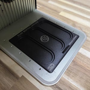 May include: A silver and black flat bottle opener tool featuring a rectangular base with a textured black center insert. The device is marked with the text FLAT BOTTLE OPENER and 89mm x 32mm, along with a circular logo and a 3D printed icon.