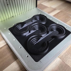 May include: A silver and black 3D printer with a series of black, semi-circular objects. The printer has a rectangular base and a vertical, ridged component. The objects appear to be molds or components for a larger project.