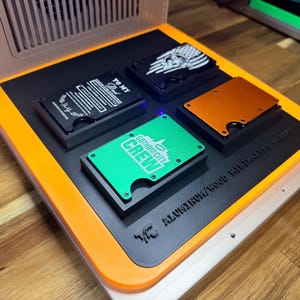 May include: Four metal wallets with various designs are displayed on a black and orange platform. One wallet is green with "GOTHAM CITY CREW" text, another has a skull and flag design, and a third has text that reads "TO MY Dad".