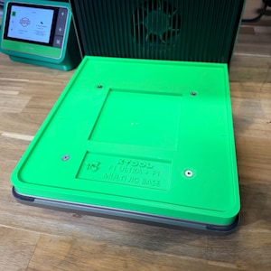 May include: A bright green XTOOL F1 Ultra Multi Jig Base, with a rectangular design and a recessed central area. The base has a dark gray border and is set on a wooden surface. The text "XTOOL F1 ULTRA + F1 MULTI JIG BASE" is visible.