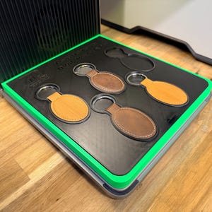 May include: A display of leather keychains in various shades of brown and tan, each with a silver keyring. The keychains are oval-shaped and set in a black tray with a green border. The tray is inside a silver case.