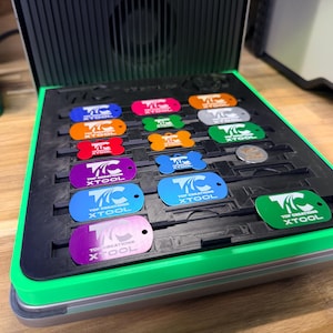May include: A tray with a green trim contains several colourful metal tags. Each tag displays the text "TOP CREATIONS XTOOL" and a logo. The tags are in shades of blue, orange, red, purple, and green.