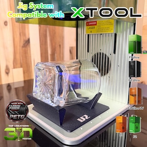 May include: A black plastic jig system for an xTool laser machine, featuring a clear glass tumbler with a starburst pattern base. The device includes a yellow laser safety warning label and text reading Jig System Compatible with xTool.