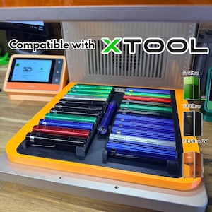 May include: An orange tray holds an assortment of colorful cylindrical objects, including black, red, green, and blue. The text "Compatible with XTOOL" is displayed above the tray. A small screen and three product images are also visible.