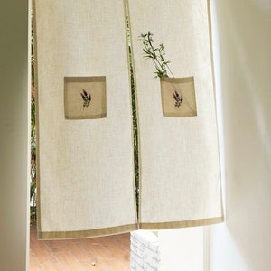 May include: Beige linen curtain panels with a brown border and square pockets. Each pocket features a lavender floral embroidery design. A sprig of greenery is placed in one of the pockets. The curtains hang in a doorway.