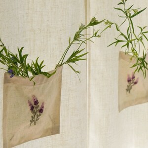 May include: Two beige fabric pockets with embroidered purple flowers hang on a textured, off-white background. Each pocket holds green foliage and small flowers, creating a natural, decorative display. The pockets are attached to a curtain or wall.