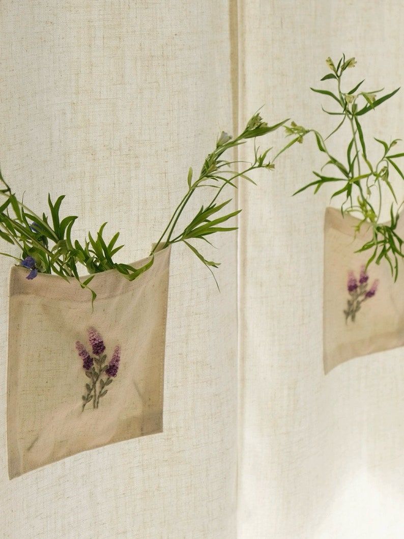 May include: Two beige fabric pockets with embroidered purple flowers hang on a textured, off-white background. Each pocket holds green foliage and small flowers, creating a natural, decorative display. The pockets are attached to a curtain or wall.