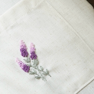 May include: Close-up of a cream-colored linen pocket with an embroidered lavender sprig. The lavender features shades of purple and pink, with green leaves and stems. The pocket is part of a larger piece of linen.