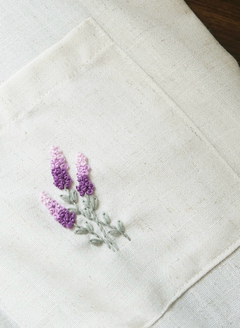 May include: Close-up of a cream-colored linen pocket with an embroidered lavender sprig. The lavender features shades of purple and pink, with green leaves and stems. The pocket is part of a larger piece of linen.
