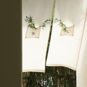 May include: Off-white linen curtains with a pocket detail holding a small bouquet of flowers. The curtains are hanging in front of a bamboo forest, creating a natural and serene aesthetic. The curtains have a light beige trim.