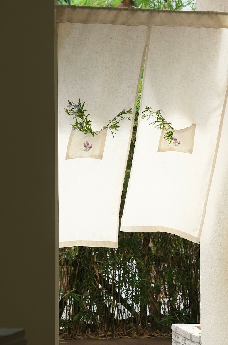 May include: Off-white linen curtains with a pocket detail holding a small bouquet of flowers. The curtains are hanging in front of a bamboo forest, creating a natural and serene aesthetic. The curtains have a light beige trim.