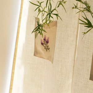 May include: A beige linen pocket planter with a lavender floral embroidery, holding green leafy plants. The planter hangs against a textured, off-white linen curtain, with sunlight casting shadows.