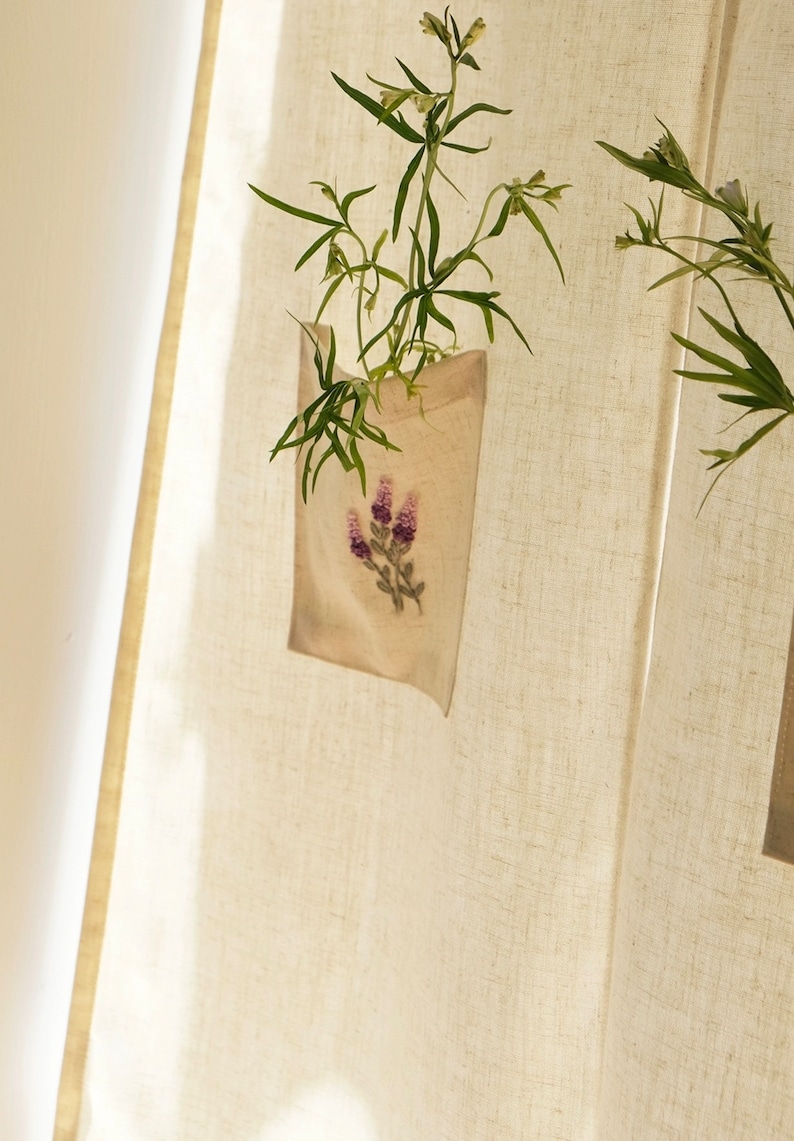 May include: A beige linen pocket planter with a lavender floral embroidery, holding green leafy plants. The planter hangs against a textured, off-white linen curtain, with sunlight casting shadows.