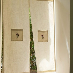 May include: Beige linen curtains with a lavender floral embroidery detail. The curtains have a rectangular patch with a lavender flower design. The edges are trimmed with a darker beige border. The curtains are hanging in front of a window.