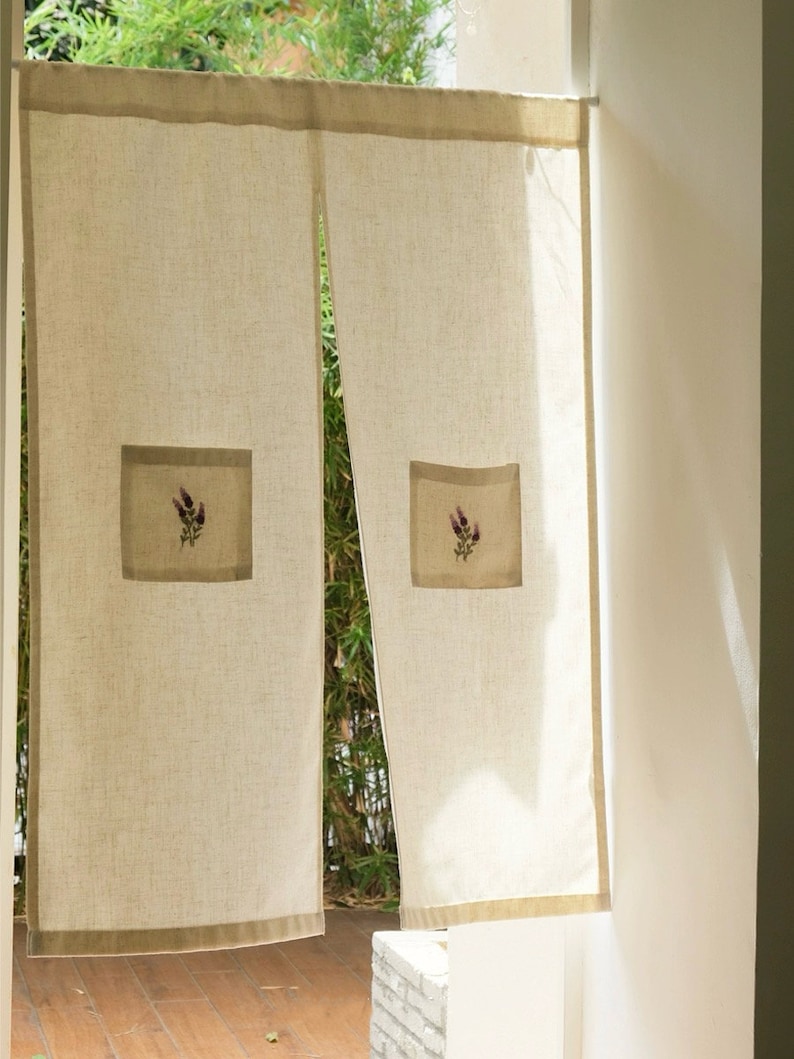 May include: Beige linen curtains with a lavender floral embroidery detail. The curtains have a rectangular patch with a lavender flower design. The edges are trimmed with a darker beige border. The curtains are hanging in front of a window.