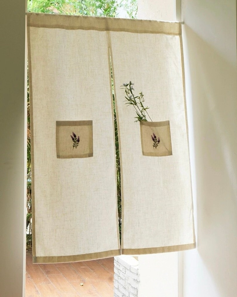 May include: Beige linen curtain panels with a brown border and square pockets. Each pocket features a lavender floral embroidery design. A sprig of greenery is placed in one of the pockets. The curtains hang in a doorway.