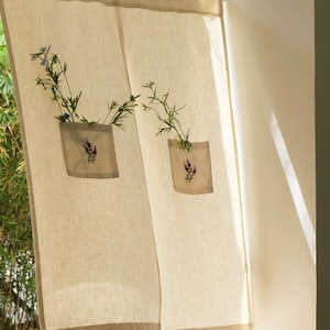 May include: Beige linen curtain with a brown border. Each panel has a square pocket with a lavender flower design and holds a small bouquet of greenery and blue flowers. The curtain hangs in front of a window.