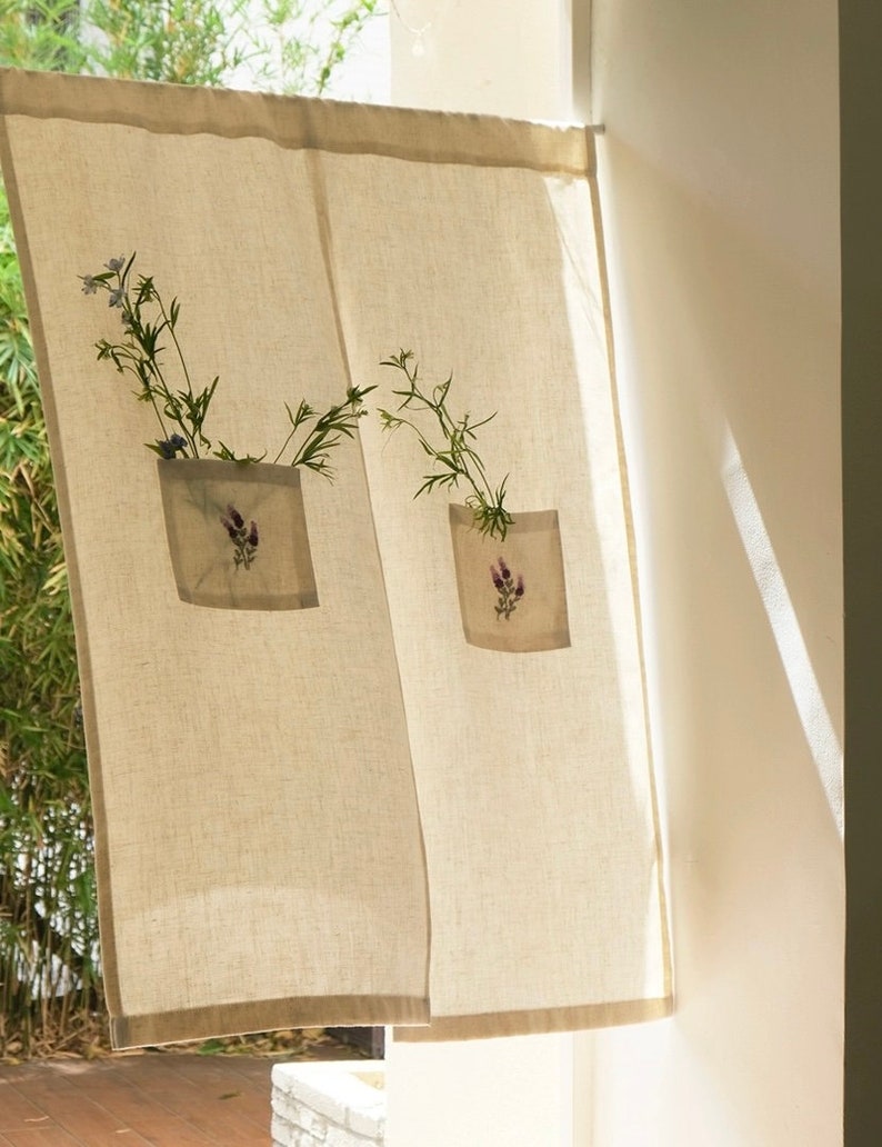 May include: Beige linen curtain with a brown border. Each panel has a square pocket with a lavender flower design and holds a small bouquet of greenery and blue flowers. The curtain hangs in front of a window.
