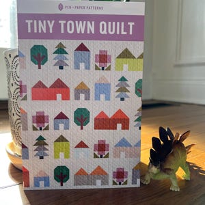 May include: A pink and white paper pattern booklet with the title "Tiny Town Quilt" and a colorful illustration of houses, trees, and flowers.