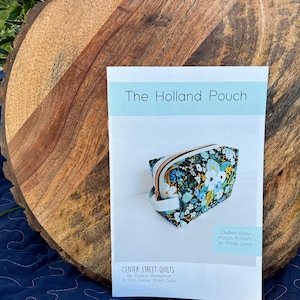 May include: A pattern for "The Holland Pouch" by Center Street Quilts, featuring a quilted boxy pouch design in three sizes. The pouch in the image has a floral pattern with a zipper closure and a small handle. The pattern is on a wooden surface.