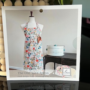 May include: A white apron with a floral print featuring red, pink, and blue flowers. The apron is on a mannequin and has a wooden spoon tucked into the pocket. The text "The One Yard Apron" and "Fresh designs from a Wyoming farm." is printed below the apron. The logo "Seun" is in the corner of the image.