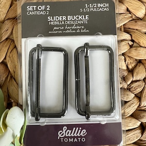 May include: A package of two black 1-1/2 inch slider buckles for purse hardware. The package is labeled "Set of 2" and "Slider Buckle".