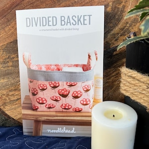 May include: A white and gray sewing pattern for a divided basket. The pattern is titled "Divided Basket" and features a pink and white fabric basket with a gray ribbon. The pattern is from Noodlehead.
