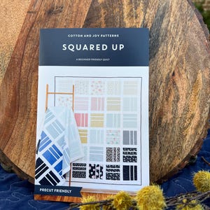 May include: A quilting pattern booklet titled "Squared Up: A Beginner Friendly Quilt" by Cotton and Joy Patterns. The cover features a colorful quilt design with a precut friendly label.