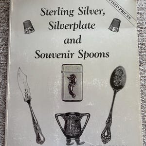 May include: A book cover titled "Sterling Silver, Silverplate and Souvenir Spoons" featuring a butter knife, lighter, spoon, thimbles, and a small trophy. The cover includes the text "Revised Prices" and "With Prices".